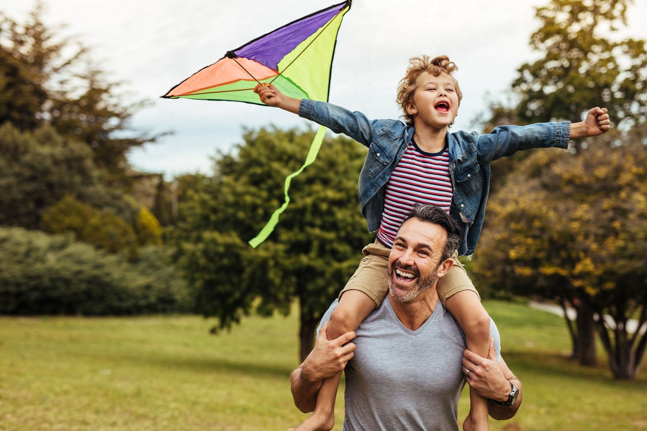 Young smiling boy playing with colorful kite outside sitting on father's shoulders. Little son with his father playing in a park with a kite.