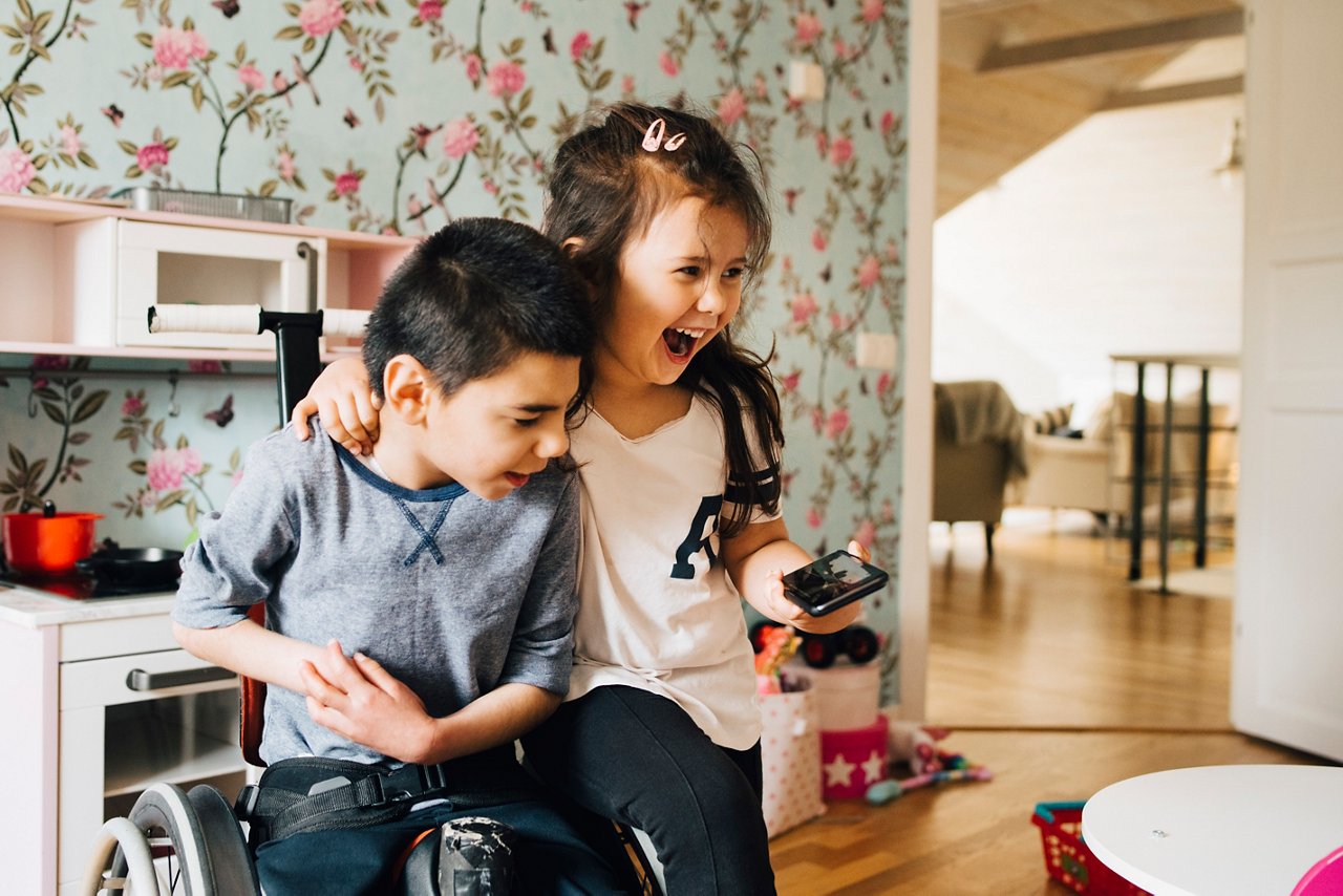 DE_nutrini_hcp_bruder_schwester_daheim_rollstuhl_schauen_auf_handy_GettyImages-1171809438