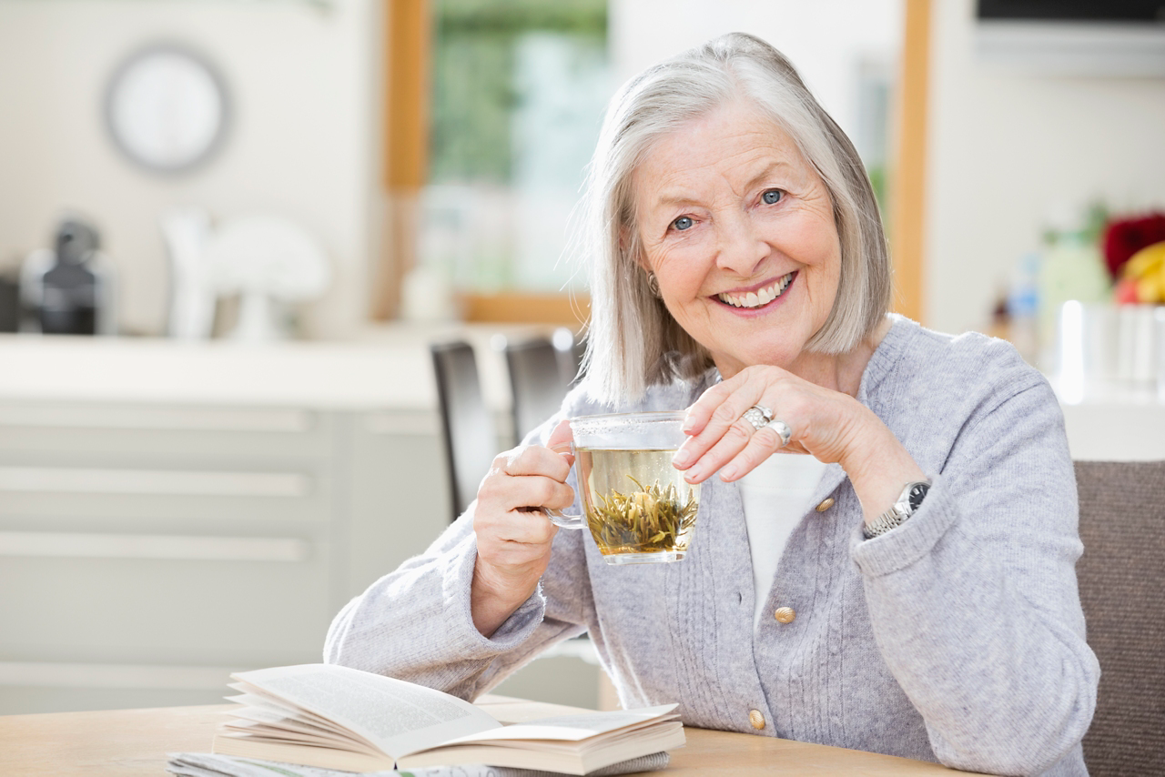 Frau mit Alzheimer-Krankheit sitzt am Tisch, trinkt Tee und lächelt