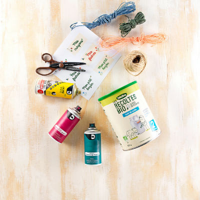 Une collection de materiel de bricolage exposee sur une surface en bois clair, comprenant des ciseaux, de la ficelle, des fils colores et des bombes de peinture. Un recipient de cereales biologiques bien visible, portant la mention � Recoltes Bio � et le chiffre � 2 �, est inclus. La scene evoque une atmosphere creative et DIY avec des couleurs et des textures vives.