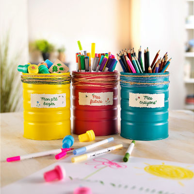 Trois bo�tes de conserve aux couleurs vives, jaune, rouge et bleu sarcelle, sont reutilisees comme rangements pour des fournitures artistiques. Chaque bo�te est etiquetee en fran�ais et contient des marqueurs, des crayons de couleur et des crayons gras. Le decor est un espace de travail lumineux et creatif, avec des fournitures artistiques eparpillees et un croquis partiellement dessine.