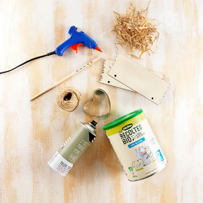 Une collection d'outils de bricolage comprenant un pistolet a colle, de la ficelle, des pieces en bois et un emporte-piece en forme de c�ur disposes sur une surface en bois clair. L'image presente un recipient pour aliments biologiques portant la mention � Recoltes Bio � avec du texte et des details numeriques visibles tels que � 2 �. Une bombe aerosol est egalement presente, renfor�ant le theme du bricolage.