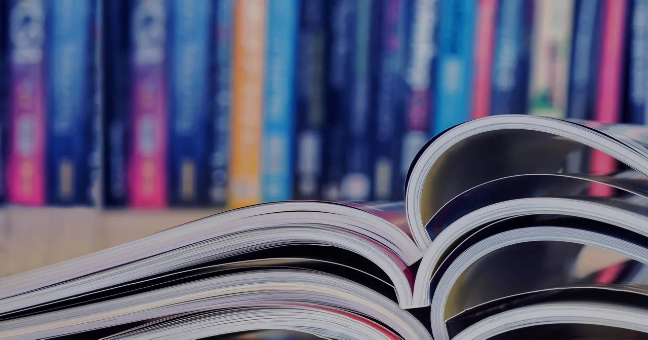 A stack of open magazines lies on a flat surface, showcasing glossy pages with vibrant visuals. In the background, a blurred array of colorful books creates a library-like setting. The image emphasizes reading materials and a creative, educational atmosphere.