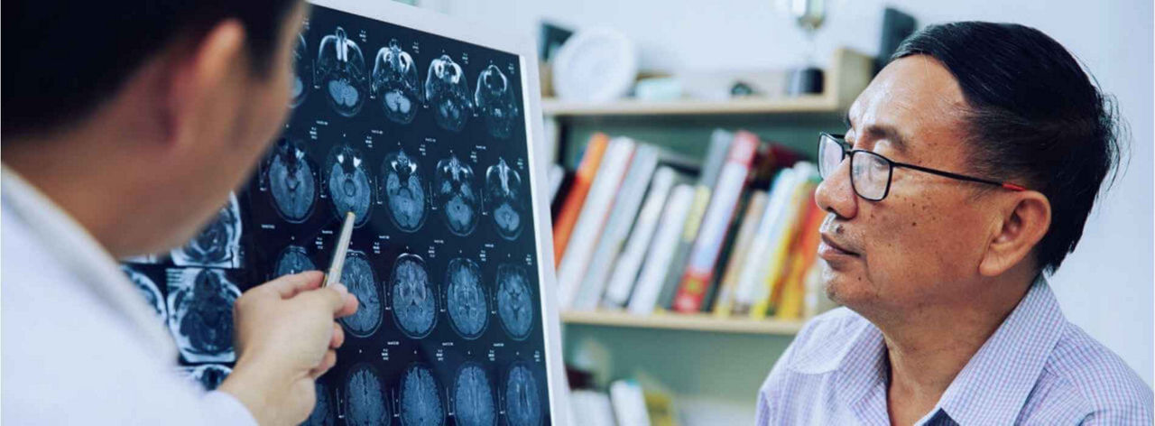 A doctor points at brain scans on a monitor, explaining results to an attentive older man. Shelves with books in the background suggest a clinical setting.