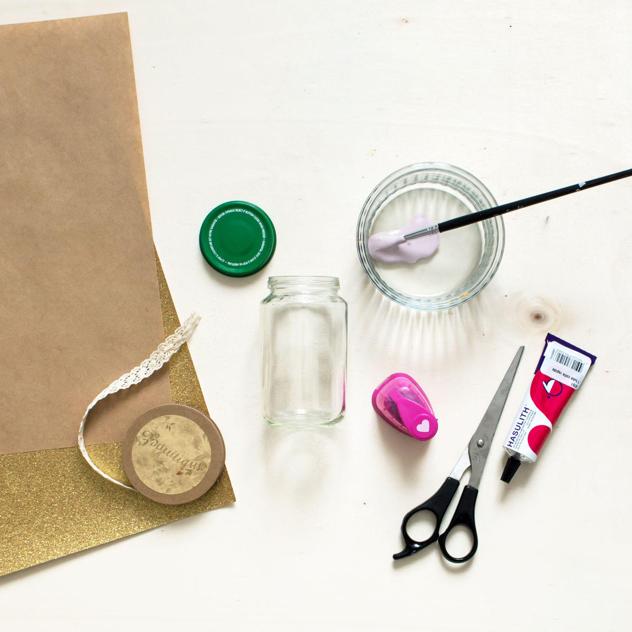 Une collection de fournitures de bricolage comprenant un bocal en verre, des ciseaux, un ruban en dentelle et un tube de peinture rose. La mise en scene comprend une feuille pailletee doree, un couvercle de bocal vert et un petit tampon en forme de c�ur. Un pinceau trempe dans de la peinture rose repose dans un bol transparent, suggerant une activite creative en cours.