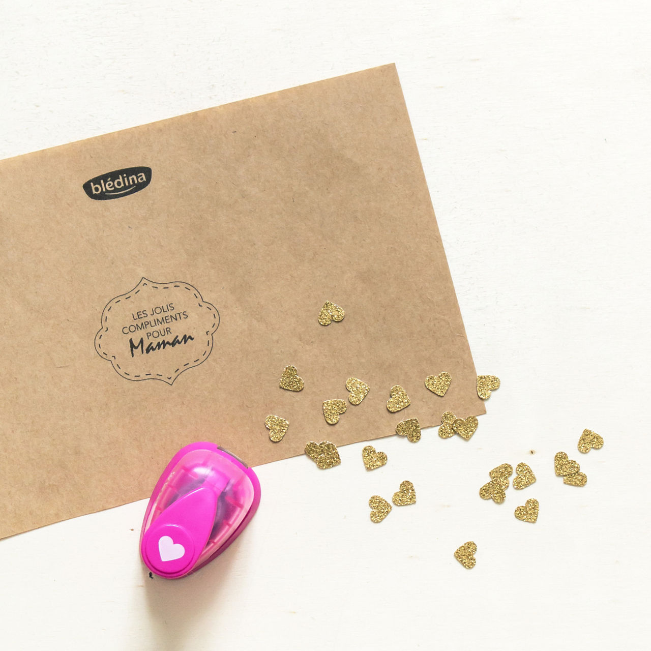 Une perforatrice rose en forme de c�ur est posee sur une surface blanche, a c�te de confettis dores en forme de c�ur eparpilles. On aper�oit une enveloppe marron sur laquelle figurent le texte � Les Jolis Compliments pour Maman � et le logo � bledina �. La scene evoque une activite creative et sincere.