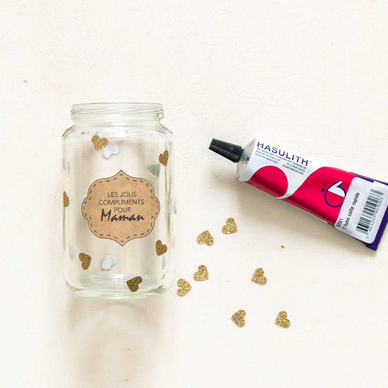 Un pot en verre transparent decore de c�urs dores et d'une etiquette sur laquelle on peut lire � Les Jolis Compliments Pour Maman �. a c�te du pot se trouve un tube de colle Hasulith, avec des motifs rouges et violets vifs. La scene se deroule sur un fond clair et neutre, parseme de c�urs dores.