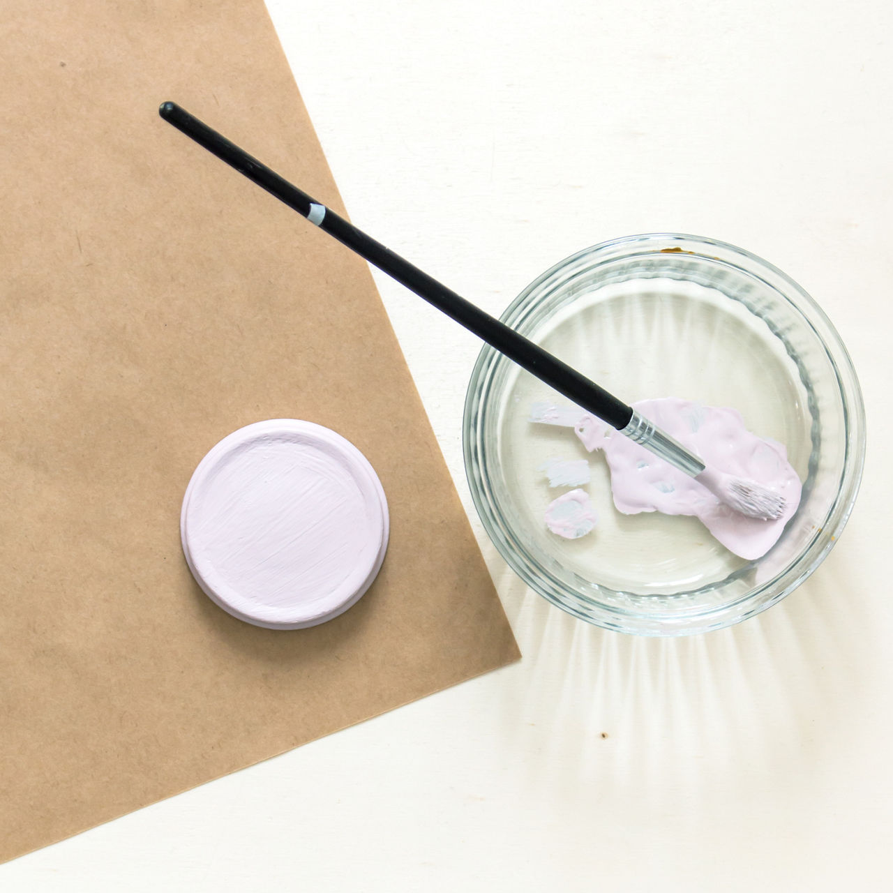 Un pinceau a manche noir repose sur un pot en verre contenant de la peinture rose pastel. La scene comprend un couvercle de pot de peinture ferme et une surface en papier brun. La palette de couleurs douces cree une atmosphere calme et creative.