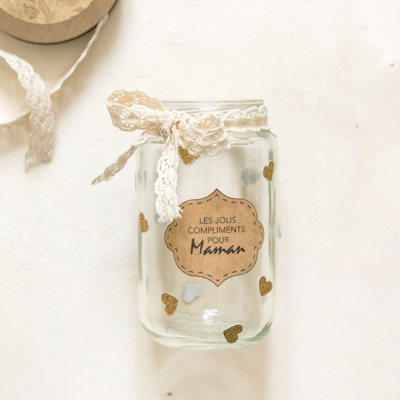 Un pot en verre orne de motifs en forme de c�ur dores et d'un ruban en dentelle, avec une etiquette sur laquelle on peut lire � Les Jolis Compliments Pour Maman �. Le pot est pose sur une surface de couleur claire, dans un style rustique et elegant. Le decor evoque une ambiance chaleureuse et sentimentale, ideale pour offrir un cadeau ou decorer.
