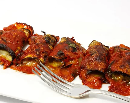 Close-up of stuffed eggplant rolls served on a white plate. The rolls are covered in a rich tomato sauce and appear baked or grilled, showcasing a rustic and appetizing texture. A silver fork is placed beside the rolls, adding a casual dining element to the composition.