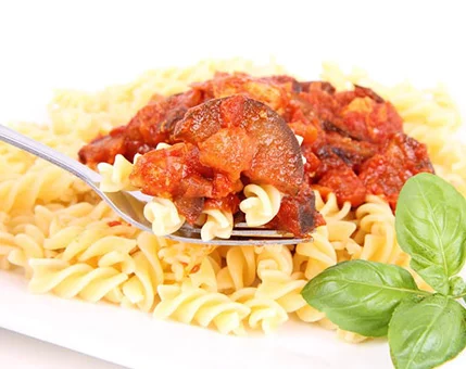 A close-up of cooked fusilli pasta topped with a rich tomato-based sauce featuring chunks of vegetables. A fork is lifting a portion of the pasta, highlighting the texture and vibrant colors. Fresh basil leaves are visible on the plate, adding a touch of green to the composition.