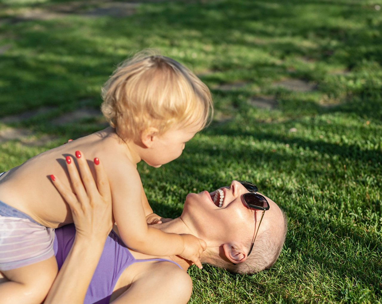 Young adult beautiful bald caucasian mom enjoy lying with little blond toddler boy son on green grass lawn at city park at summer day. Mother happy parenting and childcare . Healthy baby outdoors.