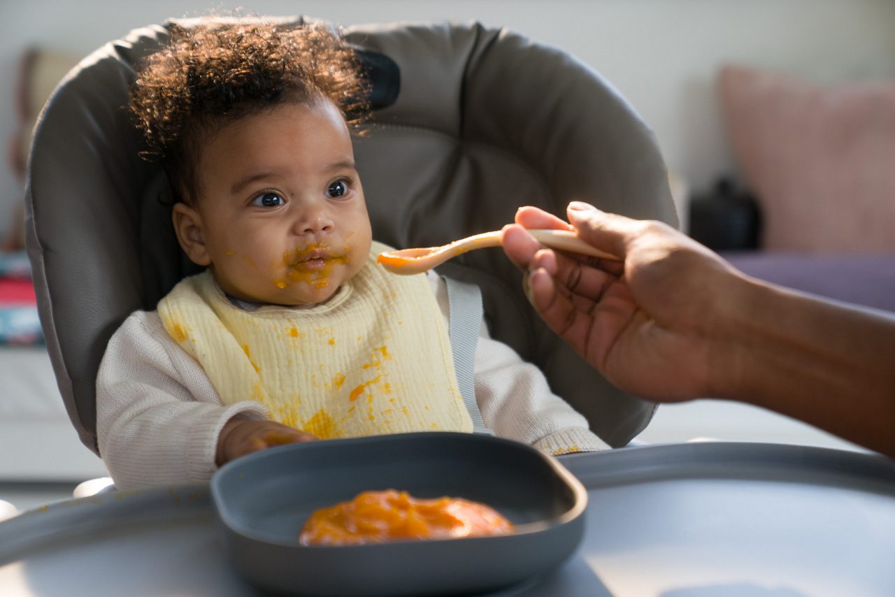 Baby in een kinderstoel die babyvoeding eet met een lepel, gemaakt met ingrediënten geschikt voor jonge kinderen.