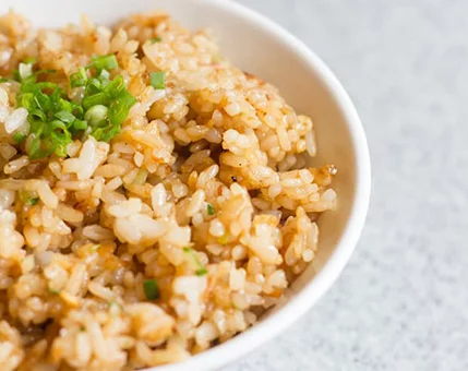 A bowl of golden-brown fried rice garnished with fresh green onions. The dish is presented on a light gray textured surface, emphasizing its appetizing appearance. The rice grains are evenly cooked, showcasing a mix of flavors and textures.