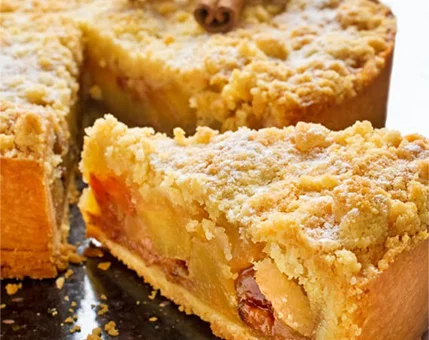 A freshly baked apple pie with a golden crumbly crust is displayed on a dark surface. The filling includes visible apple slices and raisins, complemented by a cinnamon stick garnish. The pie is dusted with powdered sugar, creating a warm and inviting presentation.