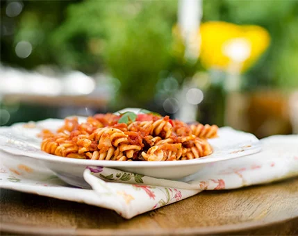 A close-up of a plate of rotini pasta topped with tomato-based sauce and garnished with fresh basil. The dish is placed on a floral-patterned napkin on a wooden table. The background features a blurred garden scene with vibrant greenery and yellow flowers, suggesting an outdoor dining experience.