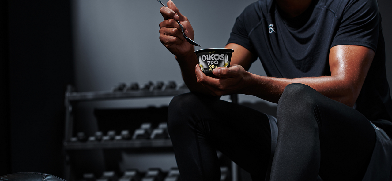 A man dressed in athletic wear is seated in a gym environment, eating a Oikos Pro yogurt cup