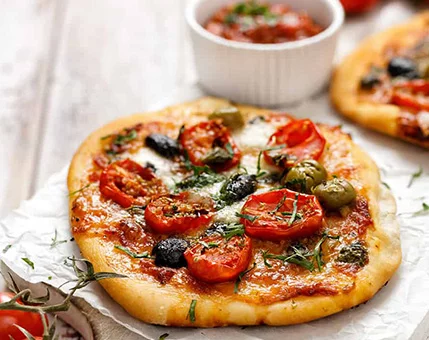 A close-up of a freshly baked pizza topped with cherry tomatoes, olives, and melted cheese, garnished with fresh herbs. The pizza is served on parchment paper with a side of tomato sauce in a white ramekin. The setting features a rustic wooden table with additional tomatoes visible in the background.
