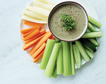 A colorful assortment of fresh vegetables, including carrots, celery, cucumber, and white root vegetables, arranged around a bowl of creamy dip. The dip is garnished with fresh herbs and pepper, adding a touch of flavor. The setting is clean and minimal, with a white marble background enhancing the vibrant colors of the vegetables.