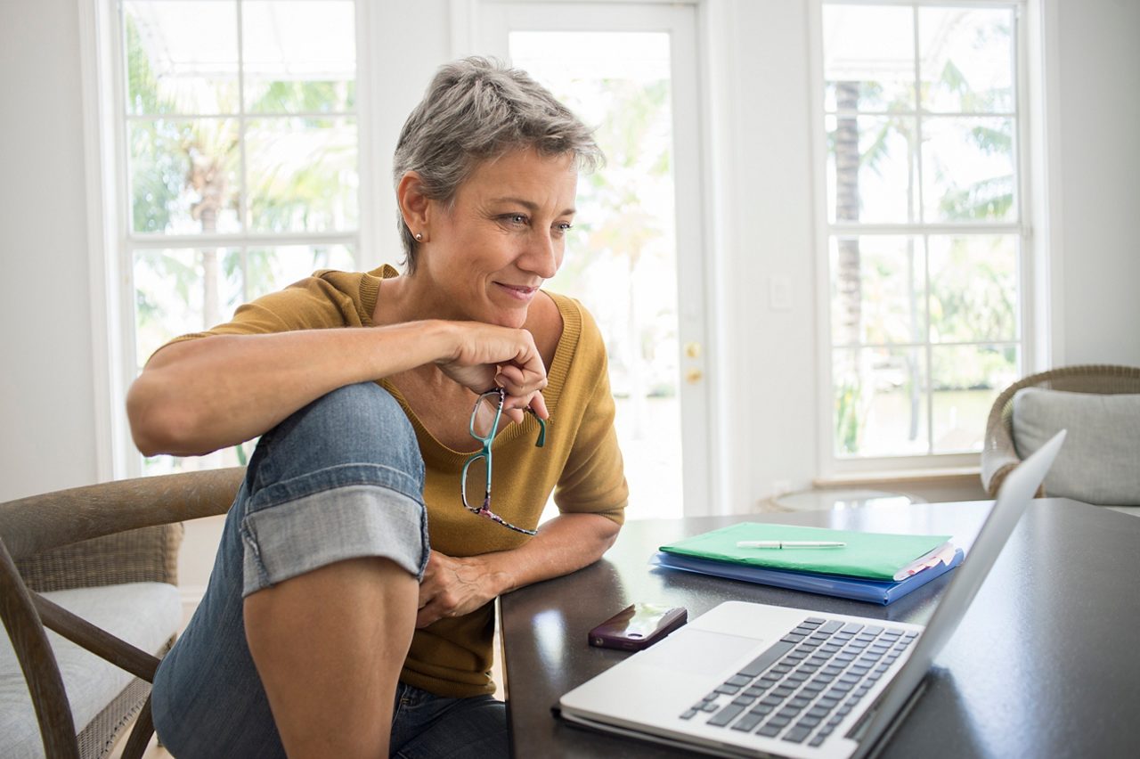 Eine Frau mit kurzen grauen Haaren sitzt vor einem Laptop am Tisch, daneben liegen Ordner, Stift und ein Handy.