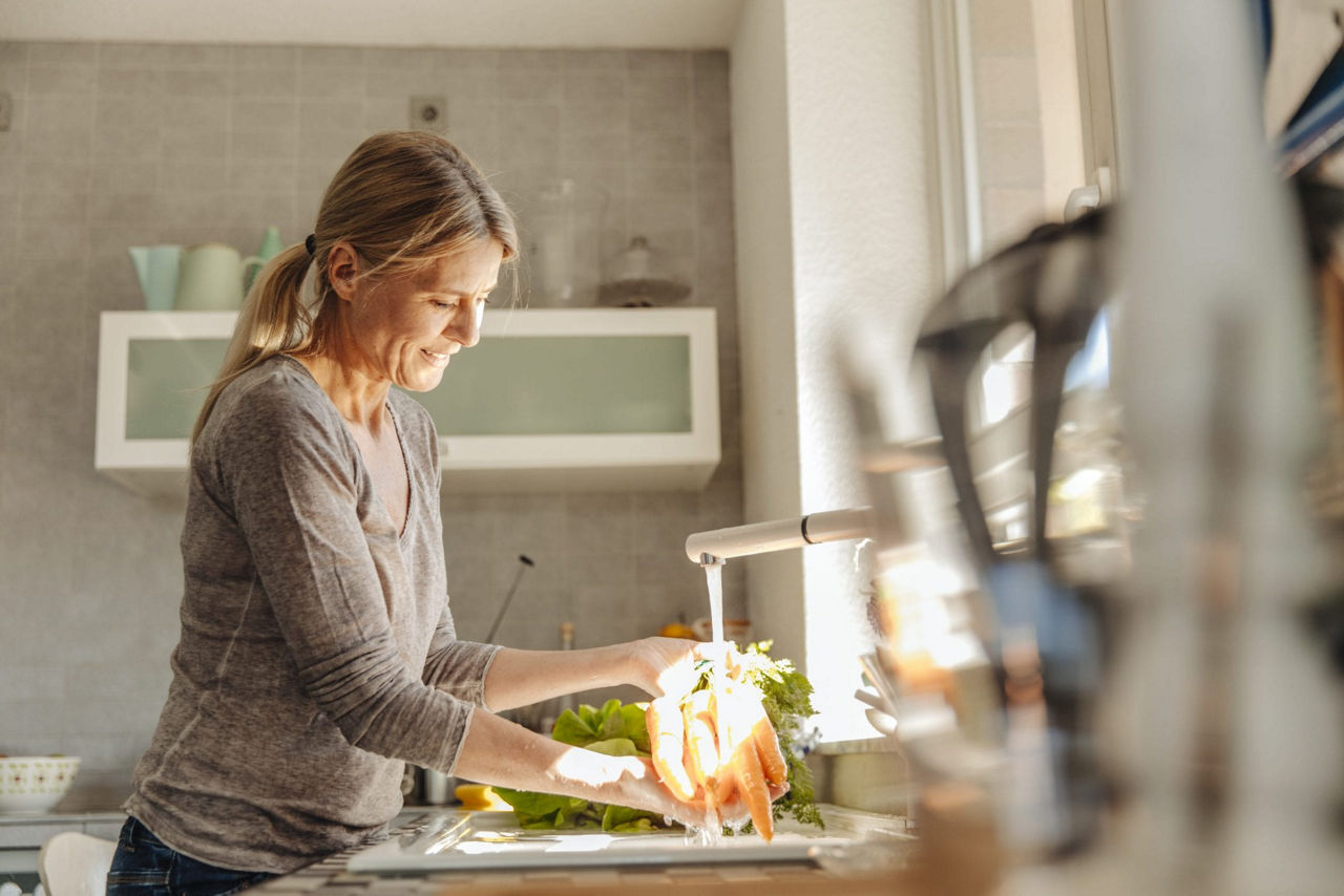 Une femme rince des legumes frais sous le robinet d'une cuisine moderne et lumineuse. Le decor comprend une lumiere naturelle qui penetre par la fenetre et une decoration minimaliste aux tons neutres. On aper�oit des legumes verts a feuilles et des carottes, soulignant un mode de vie sain et une activite de preparation des aliments.