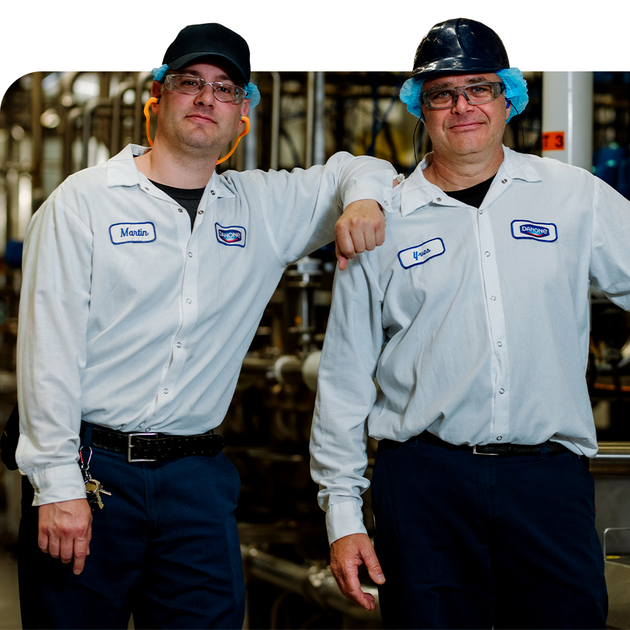 Two smiling Danone Canada employees in uniform stand arm-in-arm in a production facility.