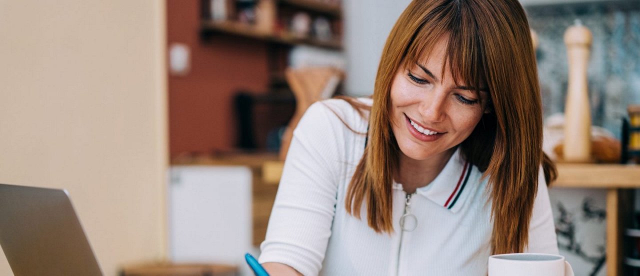 Eine Frau schreibt lächelnd an einem Tisch mit Laptop und Kaffeetasse in einer Küche mit Regalen im Hintergrund.