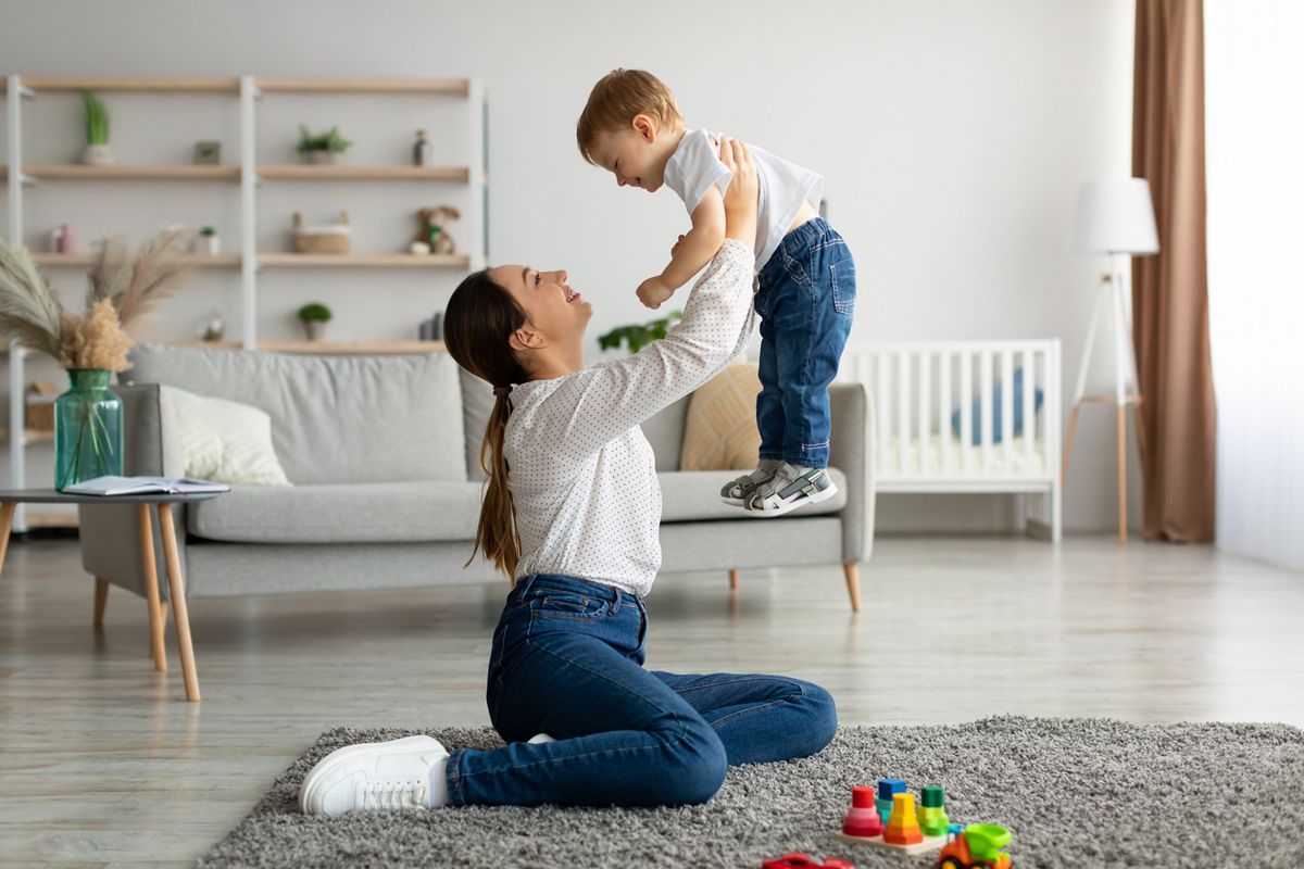 Unconditional love. Happy young mother enjoying time with her adorable toddler son at home, lifting him in the air, playing with kid boy in living room, free space