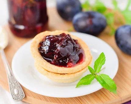 A close-up of a tart filled with rich plum jam, served on a white plate. The setting includes fresh plums and a mint leaf garnish, placed on a wooden board. A jar of plum jam and a vintage spoon are visible in the background, adding a rustic touch.