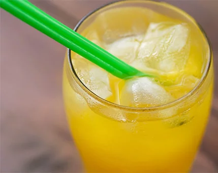 A close-up of a vibrant orange beverage served in a glass filled with ice cubes. The drink is complemented by a bright green straw, creating a visually appealing contrast. The setting suggests a casual and refreshing moment, ideal for warm weather.