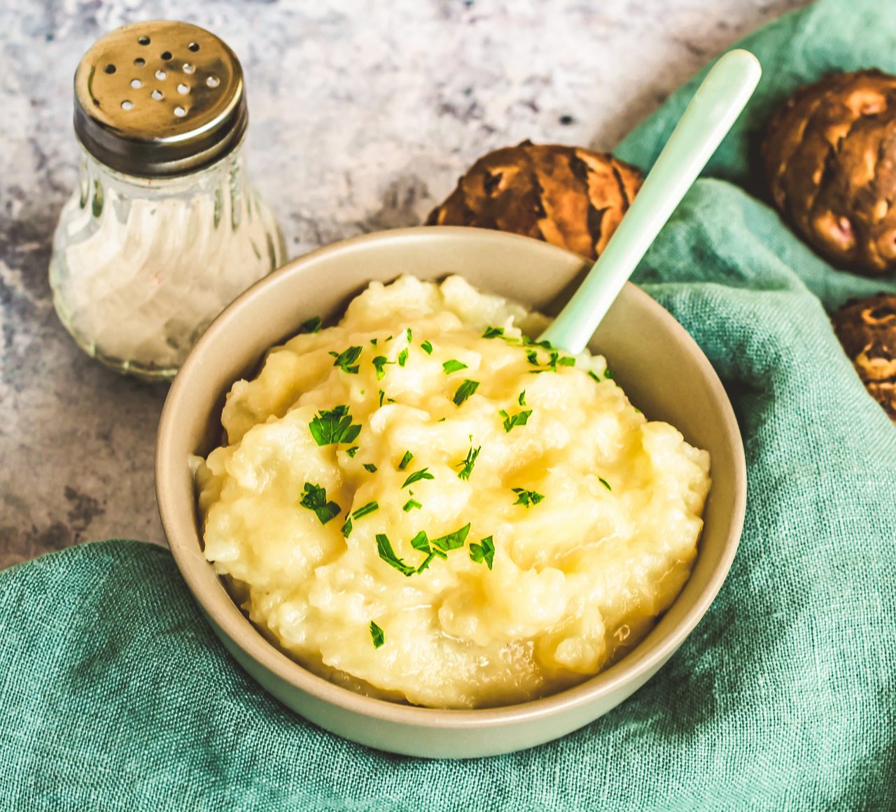 Schale mit Kartoffelpüree, bestreut mit frischer Petersilie, daneben Salzstreuer und Brötchen auf grünem Tuch.