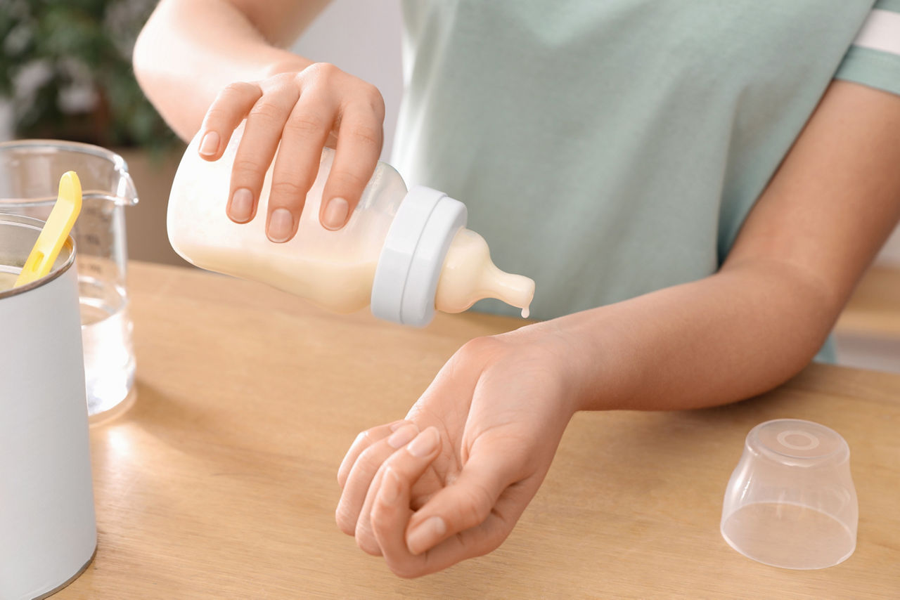 Woman checking temperature of infant formula at table indoors, closeup. Baby milk