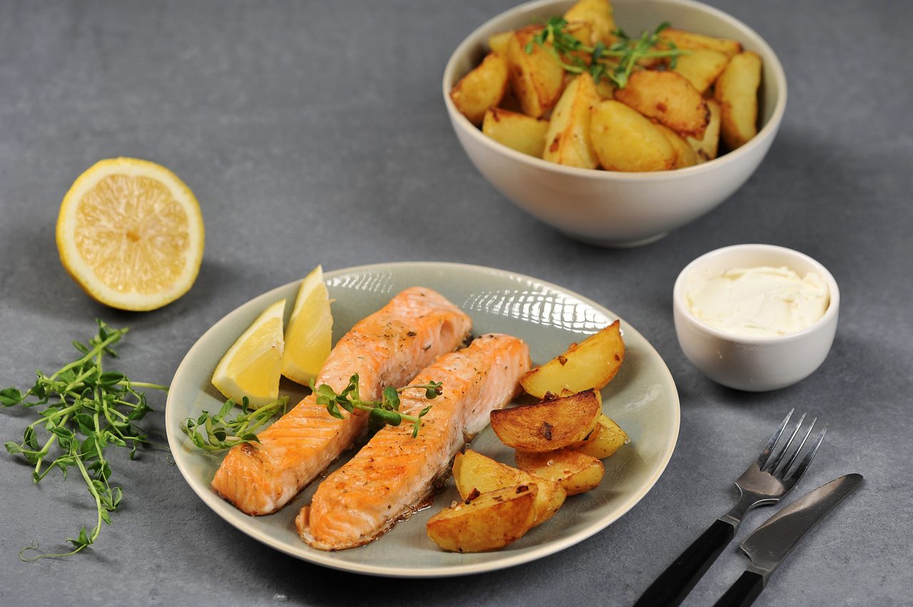 Gegrillter Lachs mit Ofenkartoffeln, Zitronenspalten, frischen Kräutern und Schälchen mit weißem Dip auf grauem Tisch.