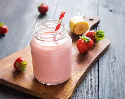 A creamy pink smoothie served in a glass jar with a striped straw, placed on a wooden board. Fresh strawberries and a sliced banana are arranged nearby, enhancing the natural and healthy vibe. The rustic wooden table adds warmth to the setting.