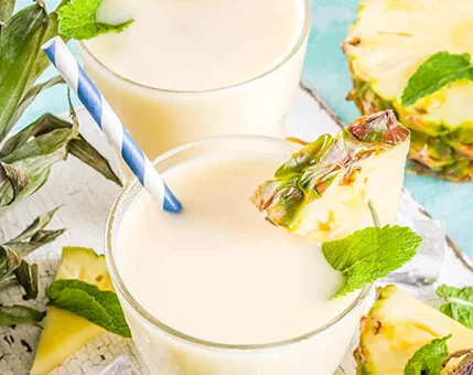 Two glasses of creamy pineapple smoothie are served with fresh mint leaves and pineapple wedges as garnish. The setting features a tropical vibe with vibrant colors and a striped straw in each glass. The drink is surrounded by pineapple slices and mint sprigs, enhancing its fresh and summery appeal.