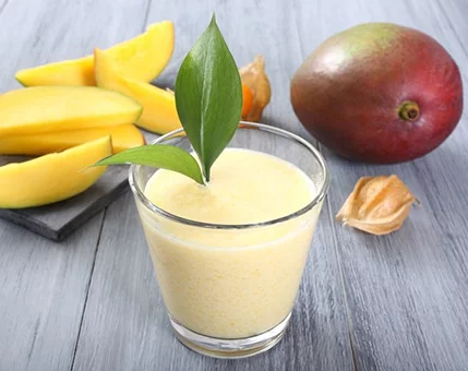 A glass of creamy mango smoothie garnished with a green leaf sits on a rustic wooden surface. Surrounding the glass are fresh mango slices, a whole mango, and golden physalis fruits, creating a vibrant tropical setting. The composition highlights the freshness and natural appeal of the ingredients.