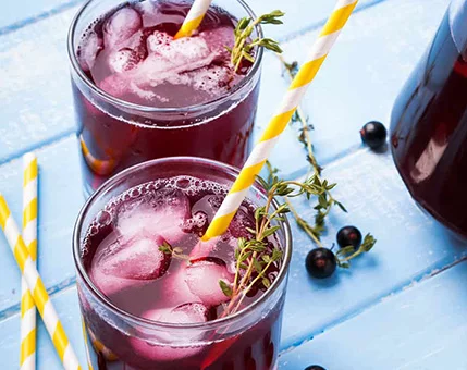 Two glasses of a vibrant purple berry beverage are placed on a light blue wooden surface. The drinks are garnished with fresh thyme sprigs and served with yellow-striped straws. Ice cubes and blackcurrants add a cooling and fruity touch to the composition.