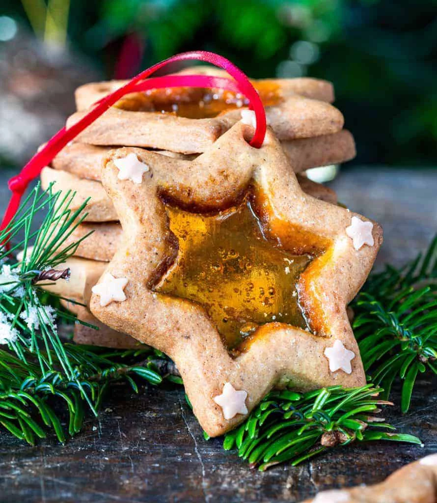 A close-up of star-shaped cookies featuring a stained glass center made of caramelized sugar. The cookies are decorated with small white star accents and tied with a red ribbon, suggesting holiday ornamentation. Set against a festive background with pine branches, the image evokes a warm and celebratory mood.