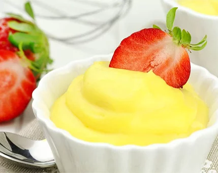 A smooth yellow dessert served in a white ceramic bowl, garnished with a sliced fresh strawberry. The setting includes additional whole strawberries and a whisk in the background, suggesting a homemade preparation. The vibrant colors create a cheerful and appetizing visual.