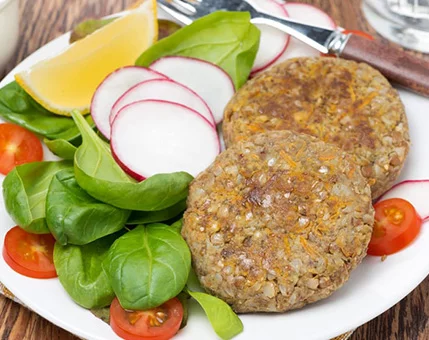 Two vegetarian patties are served on a white plate alongside fresh spinach leaves, sliced radishes, cherry tomatoes, and a lemon wedge. The setting features a rustic wooden table with a fork and knife placed nearby. The dish highlights a healthy and colorful presentation, ideal for a wholesome meal.