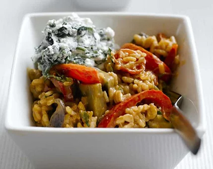 A square white bowl filled with a colorful vegetable rice dish, featuring red peppers, eggplant, and herbs. The dish is topped with a creamy spinach-based garnish, adding texture and flavor. The setting is minimalistic, with a clean white background emphasizing the food presentation.