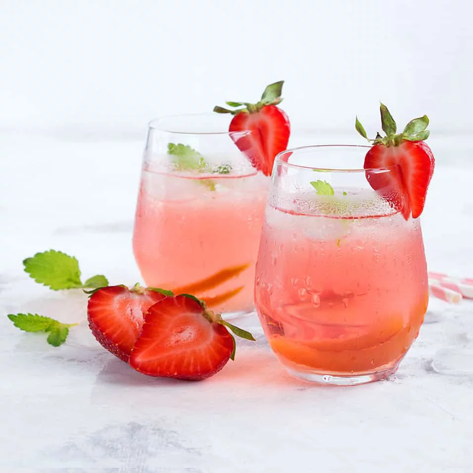 Two clear glasses filled with a pink strawberry-infused beverage are garnished with fresh mint leaves and sliced strawberries. The setting is bright and minimal, with a white background that enhances the vibrant colors of the drink. The composition evokes a fresh and summery mood, perfect for casual gatherings or relaxing moments.