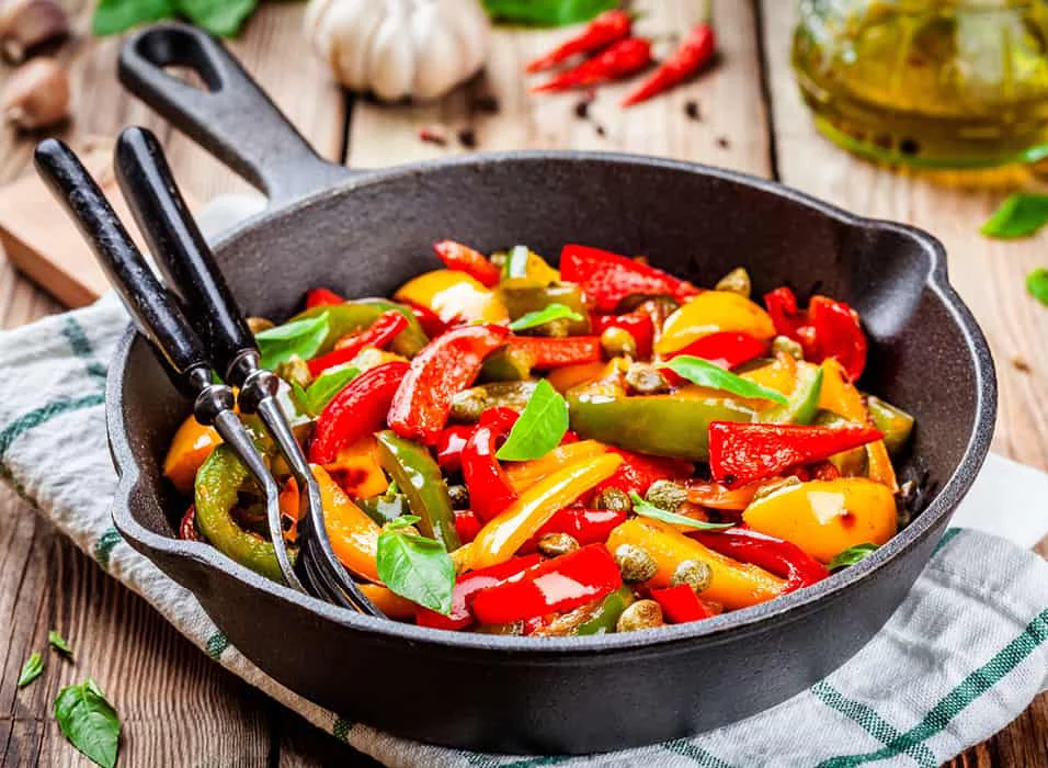 A vibrant mix of roasted bell peppers, tomatoes, and capers served in a black cast iron skillet. The setting features a rustic wooden table with fresh basil leaves, garlic, and olive oil in the background. The dish is complemented by a striped cloth and utensils placed inside the skillet.