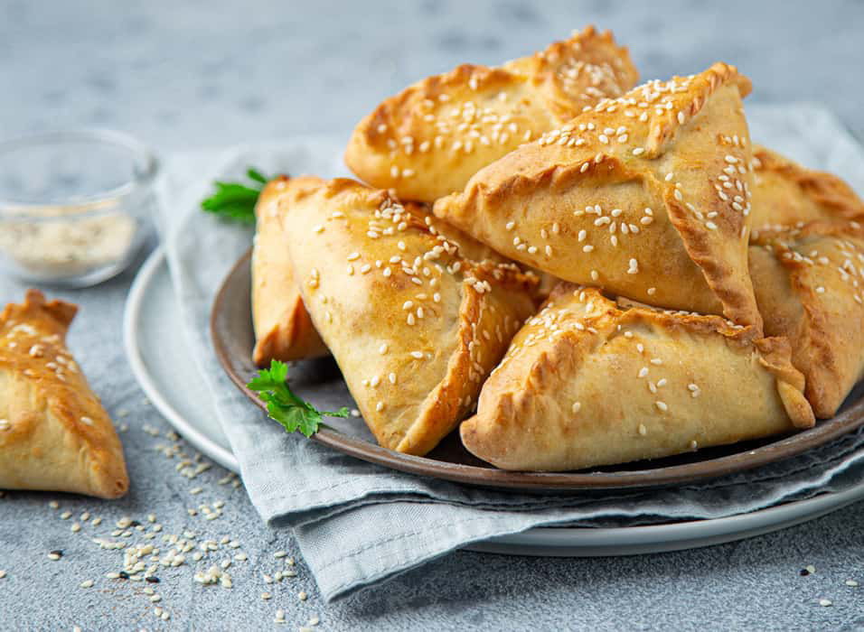 A plate of freshly baked triangular pastries topped with sesame seeds is displayed on a textured gray surface. The setting includes a small bowl of sesame seeds and sprigs of parsley for garnish. The pastries have a golden-brown crust, suggesting a warm and inviting mood.