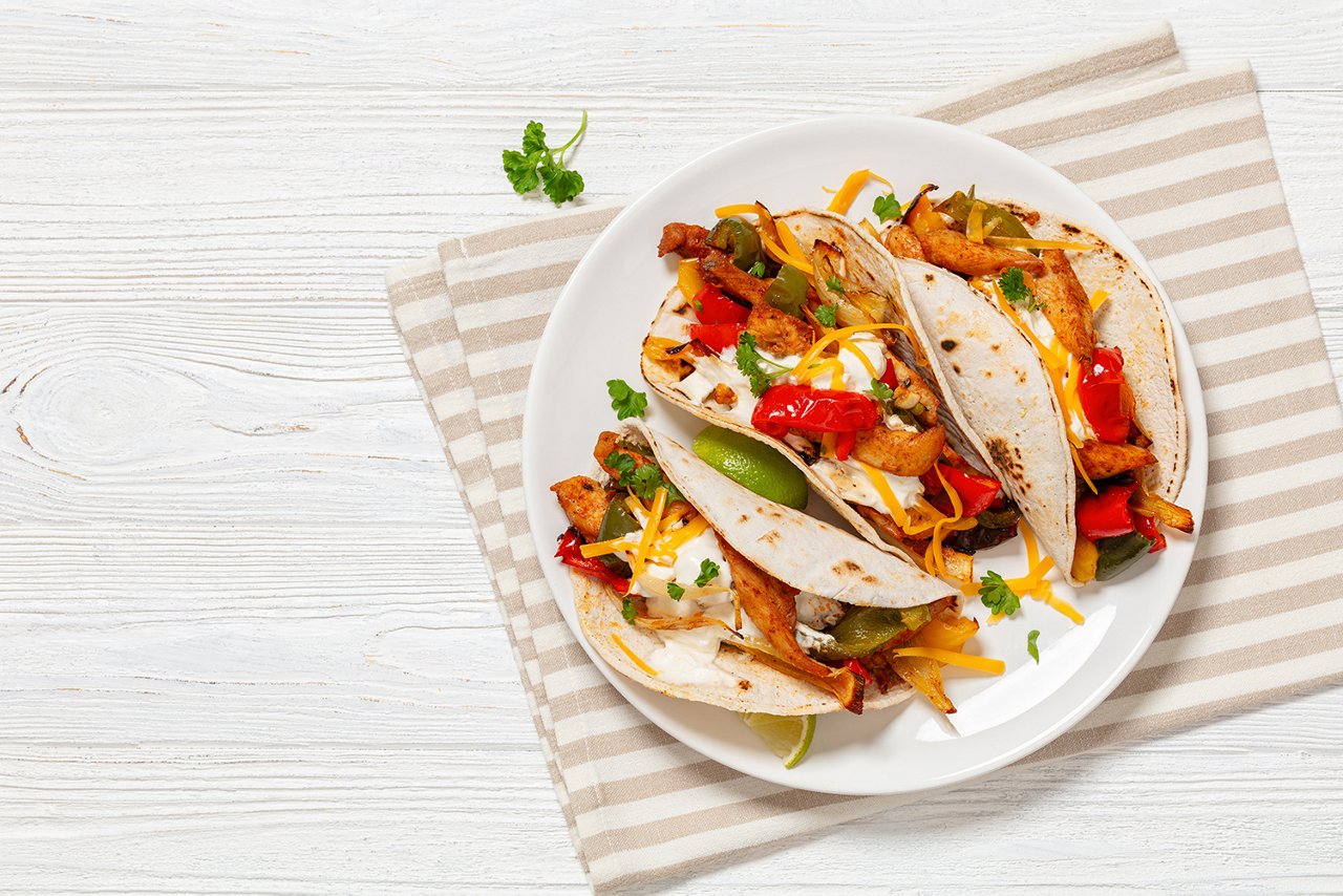 baked tex-mex chicken fajitas with mixed sweet pepper, onion, sour cream, cheddar cheese, lime, parsley and white corn tortillas on plate on white wooden table, free space, flat lay