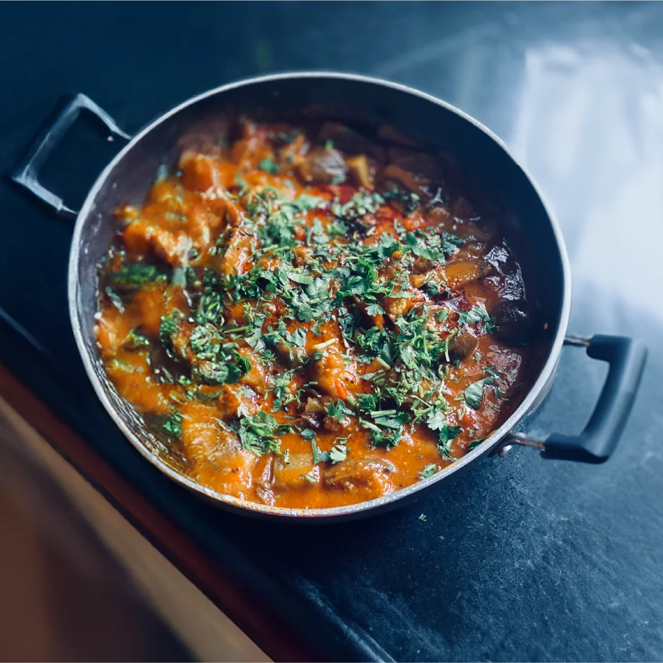 A round metal pan filled with a hearty stew sits on a dark countertop. The dish appears rich and saucy, with visible chunks of vegetables or meat in a thick orange-red gravy. Fresh green herbs are sprinkled generously on top, adding color and texture. Soft natural light highlights the steam and gloss of the simmered sauce, creating a warm, home-cooked feel.