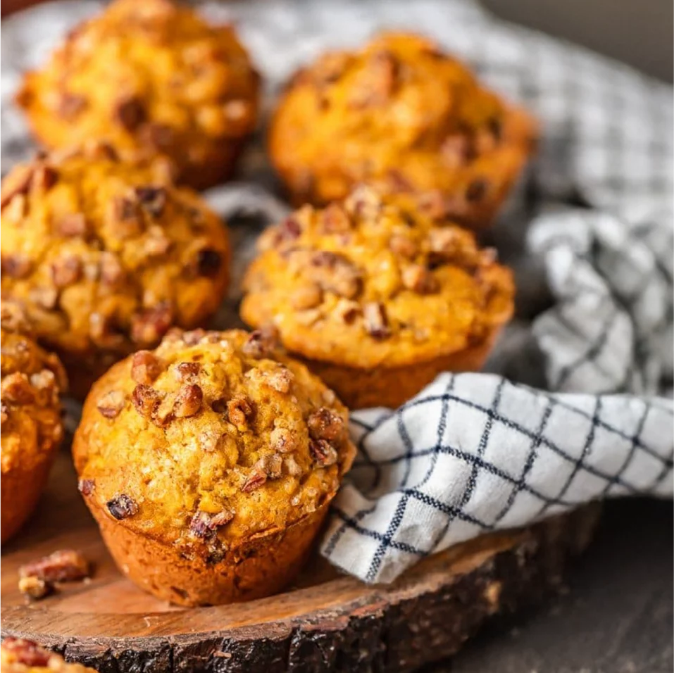 A close-up view shows several golden-brown muffins topped with chopped nuts arranged on a rustic wooden board. A checkered kitchen towel is draped around the muffins, adding a cozy, homemade feel. The background is softly blurred, keeping focus on the texture and warm color of the baked goods. The scene suggests a comforting, freshly baked treat ready to be served.