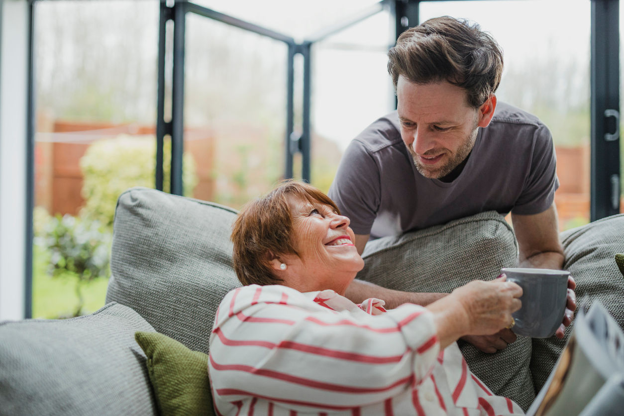 Man is leaning over the sofa to give his mother a cup of tea. She is sitting on the sofa and is taking it from him gratefully.