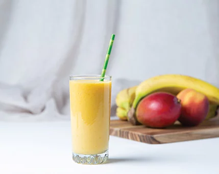 A glass of vibrant mango smoothie with a green-striped straw sits on a white surface. Behind it, a wooden tray holds ripe bananas, mangoes, and apples, creating a fresh and tropical setting. The background features soft, neutral fabric, enhancing the natural and wholesome mood.