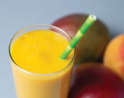 A vibrant mango smoothie served in a clear glass with a green straw. The drink is surrounded by whole mangoes and peaches, creating a colorful and fresh setting. The smooth texture and bright yellow-orange hue highlight its tropical appeal.